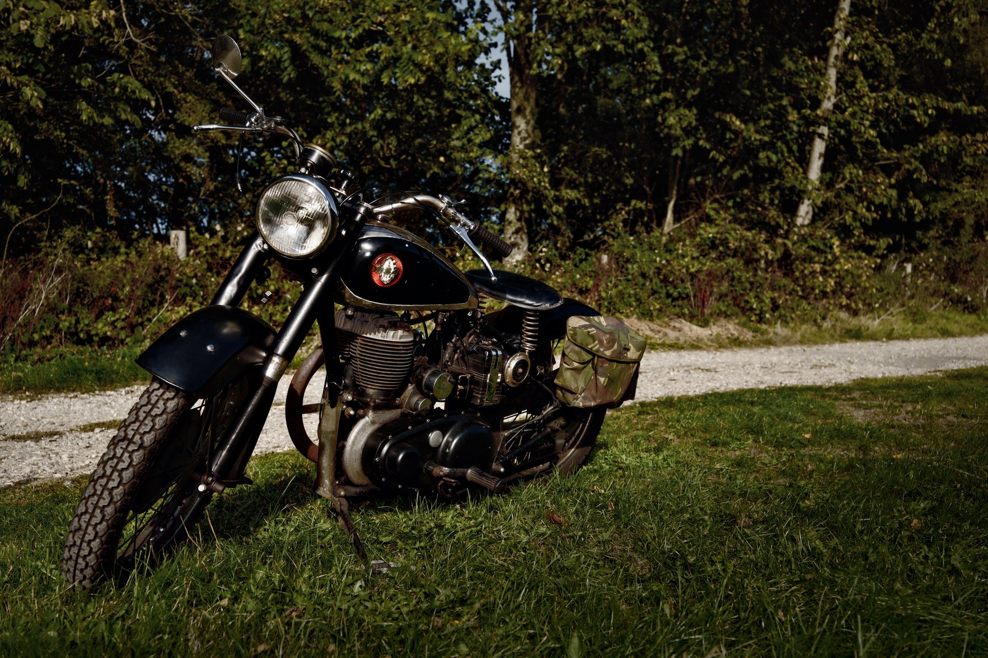 A vintage black motorcycle sits parked on green grass beside a gravel path, with dense trees in the background.