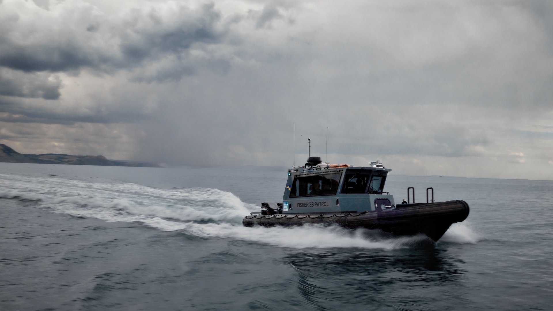 A fisheries patrol boat speeds across a gray, choppy sea beneath a heavy, cloud-filled sky with distant coastline on the horizon.