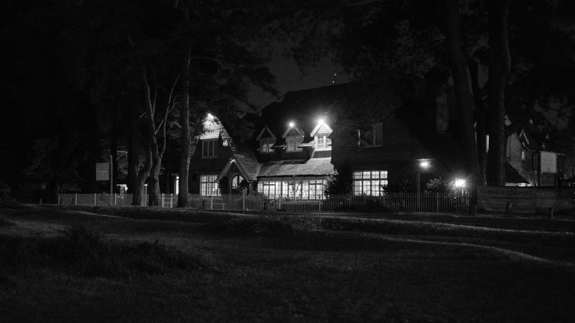 A black and white nighttime photograph of a brightly lit house with glowing windows, surrounded by dark trees and a dimly lit road in the foreground.