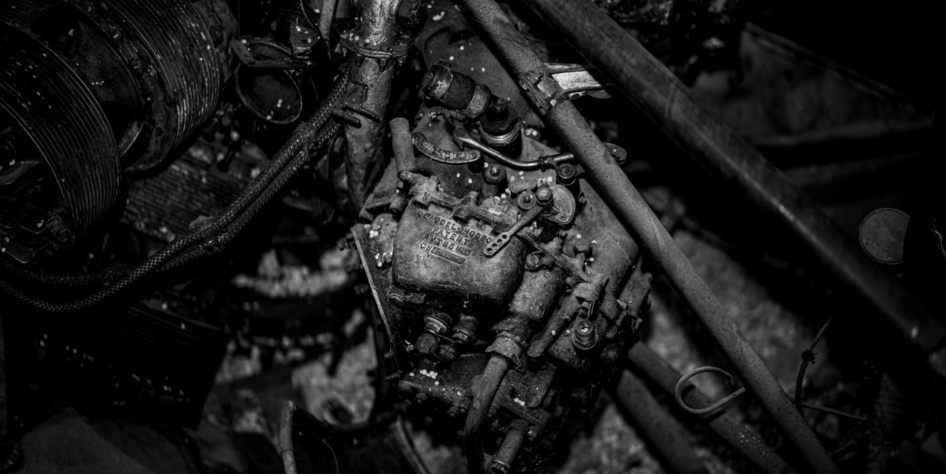 A close-up, black-and-white photograph of a corroded vintage engine covered in grime, with ribbed belts, hoses, and a faded metal nameplate at its center.