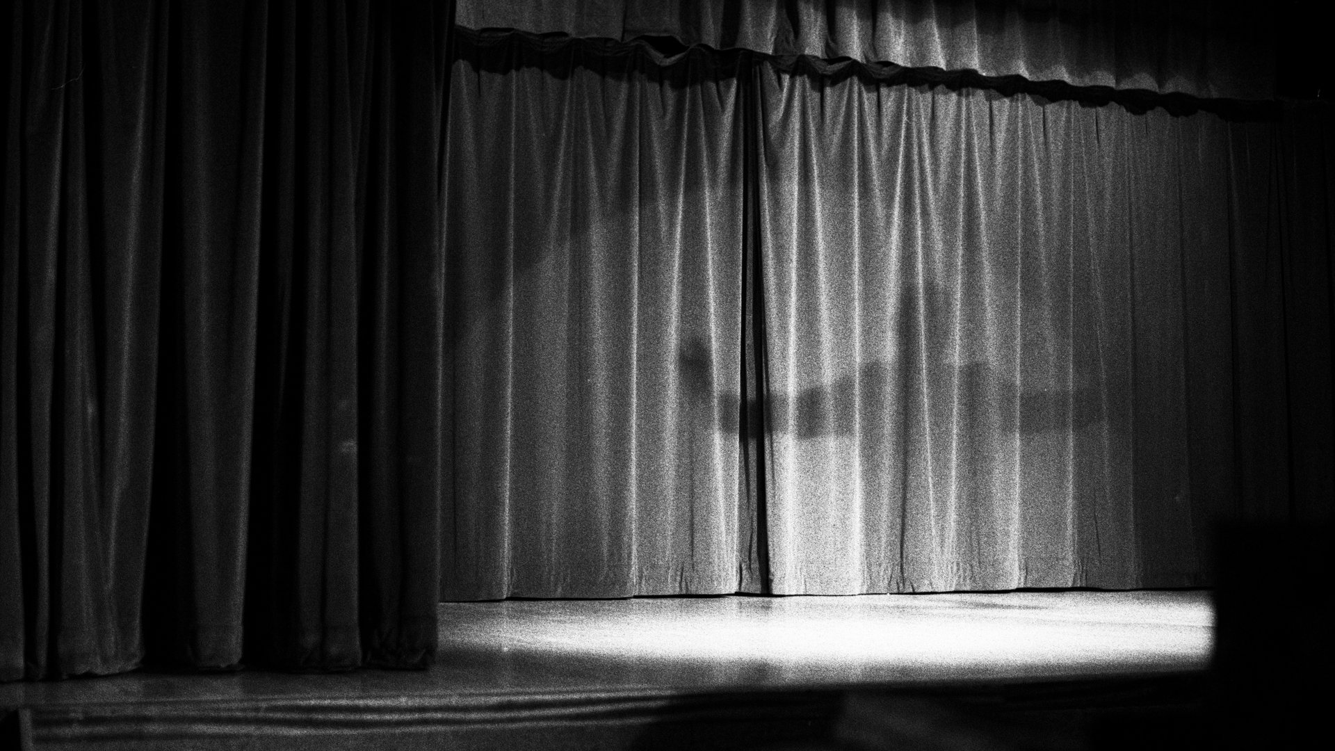 A black and white photograph of sheer curtains illuminated by window light, casting dramatic vertical shadows across the fabric and a bright window seat below.