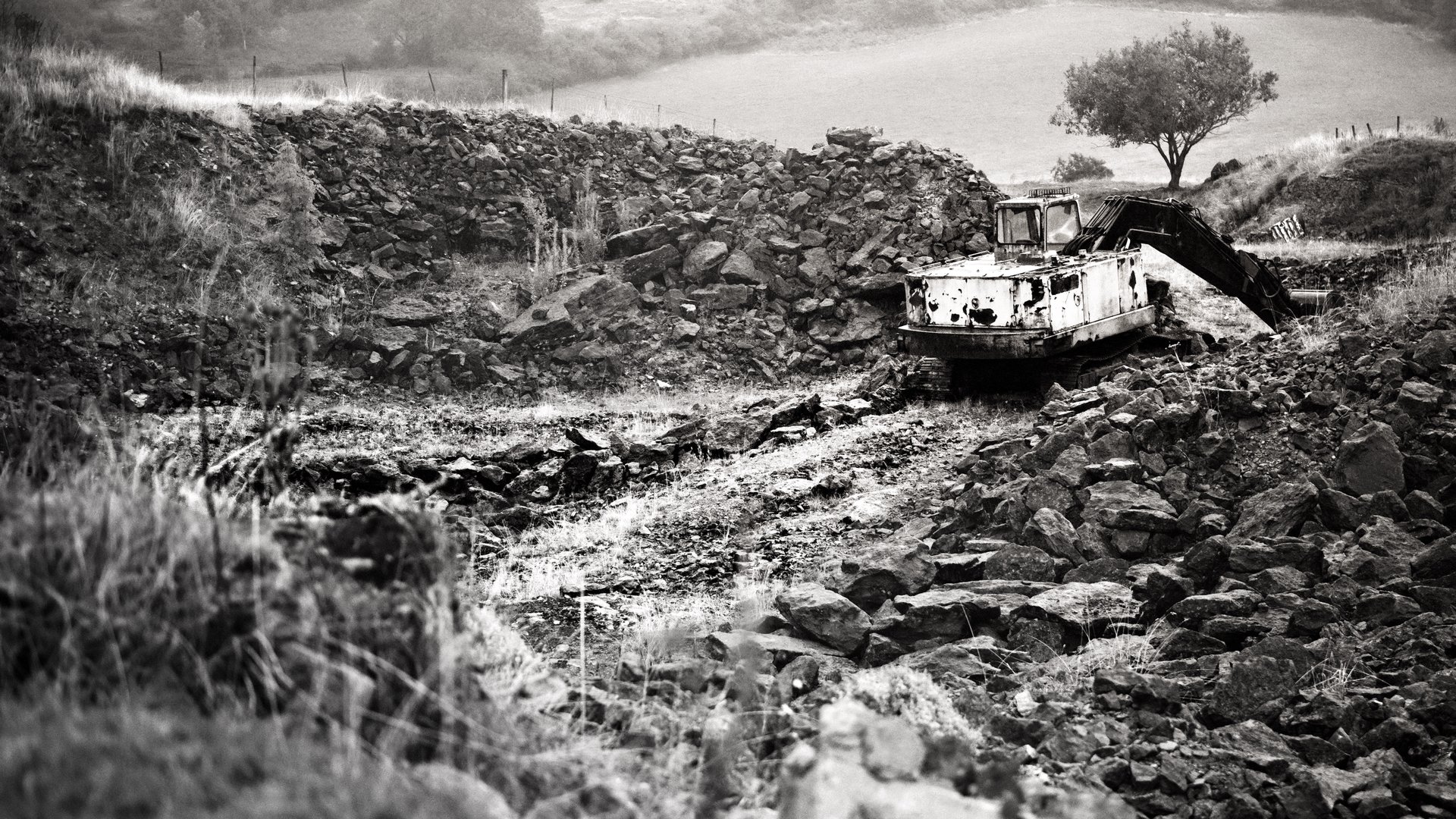 A black-and-white photograph of a rusted, abandoned excavator sitting amid jagged rocks and broken stone walls in a desolate, hilly landscape with a lone tree in the distance.